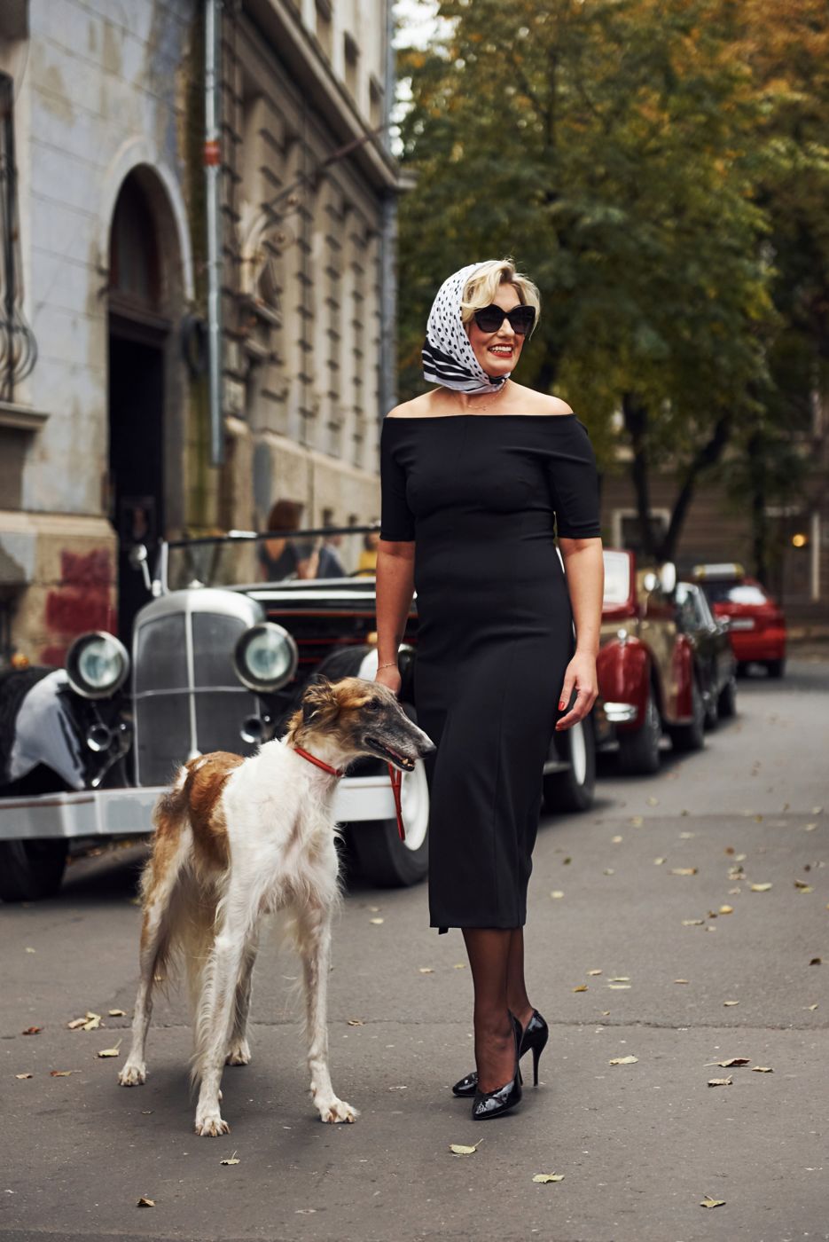 Woman in a black dress with sunglasses and a small dog beside a vintage car
